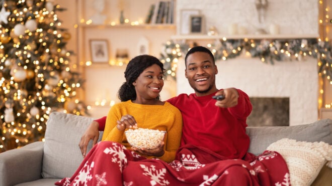 Tv programs and films at new years night at home. Young smiling african american guy switches channels with remote control, lady holds popcorn, couple covered with blanket in interior with Xmas tree