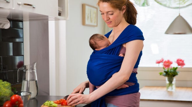 Woman with baby in a wrap carrier cooking in the kitchen, cutting vegetables with knife on a chopping board. Interior of a cozy modern kitchen. Mother doing her chores.