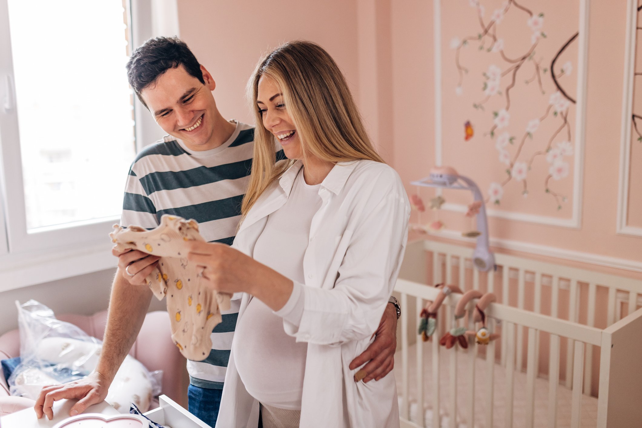 Smiling parents-to-be are standing in the baby's room and admiring the wardrobe for the baby