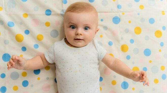 White baby wide-eyed laying on colorful sheet.