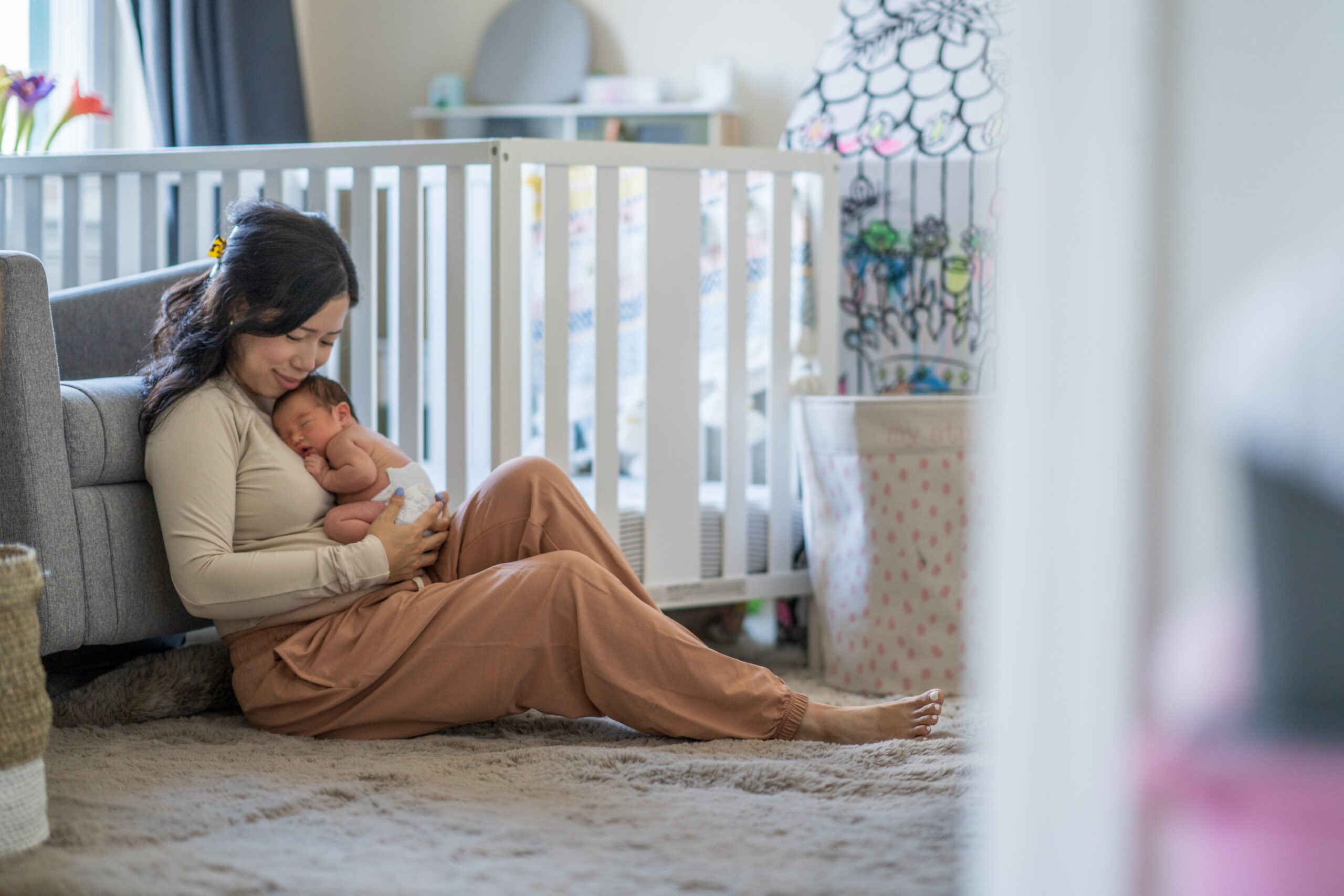 A new Mother sits on the floor of her daughters nursery as she takes a moment to enjoy Motherhood. She is dressed comfortably and holding her baby close as the two bond.