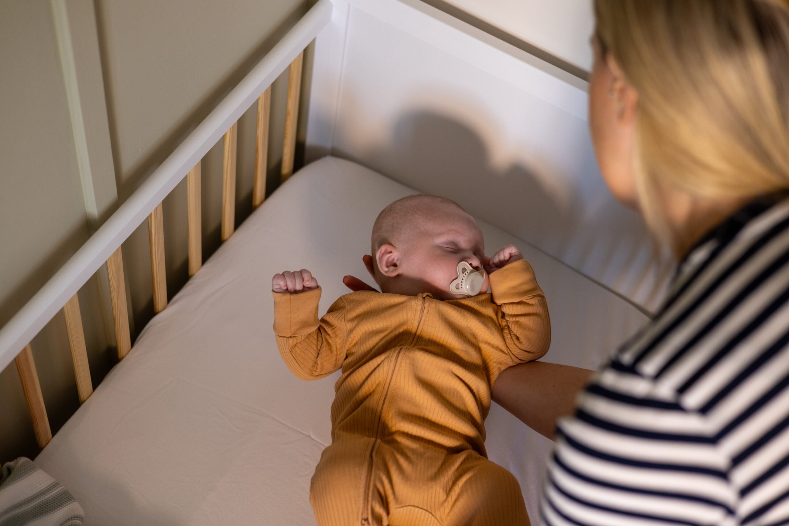 Close-up over-the-shoulder shot of a mother holding her young baby boy as she begins to place him in his cot. The woman is wearing a striped top. The baby is sleeping with a dummy in his mouth.