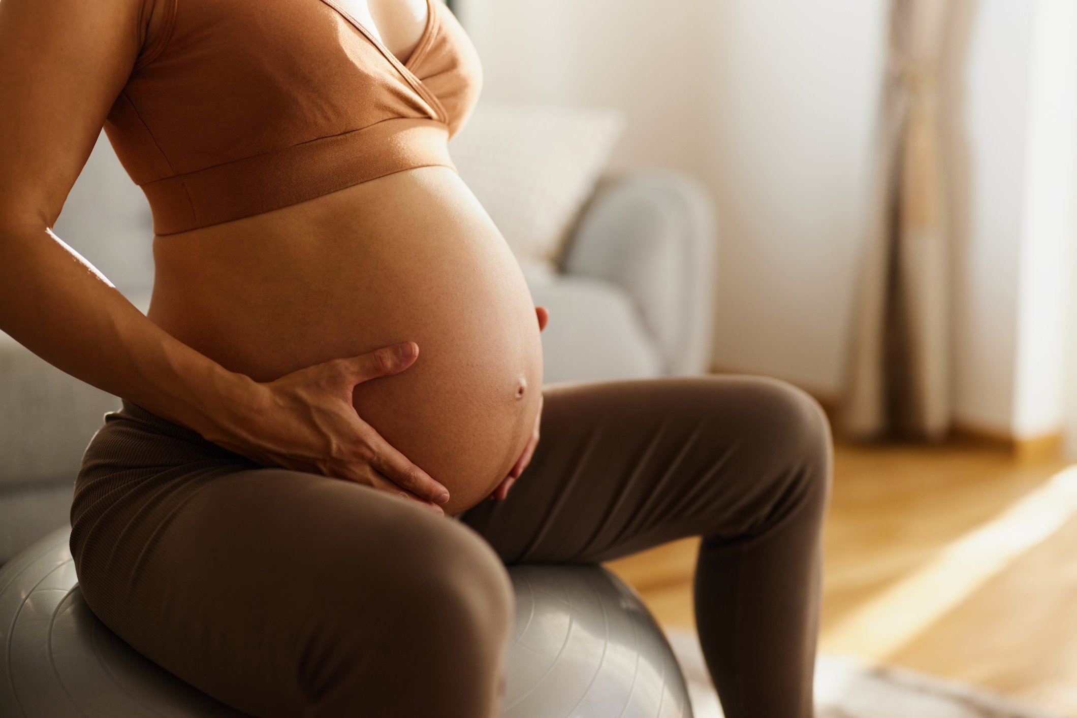 Close up of unrecognizable expecting woman holding hands on her stomach while relaxing on a fitness ball in the living room.